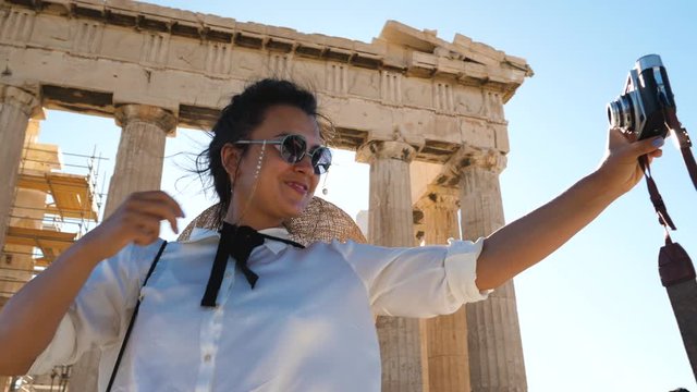 Young Woman taking selfies at Parthenon, Acropolis of Athens, Greece. Large hat, fashion white dress, sunglasses, vintage camera.