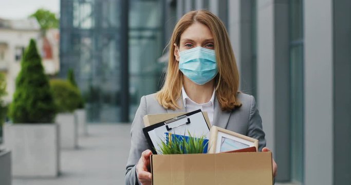 Portrait of Caucasian sad businesswoman in medical mask standing outdoor with box of stuff. Leaving business. Female office worker lost her job. Unemployment rate growing. Fired upset desperate woman.