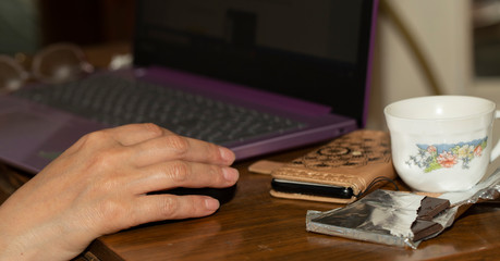Fototapeta na wymiar Self-isolation. Working remotely. A woman's hand with a mouse, laptop, coffee mug, phone and chocolate.