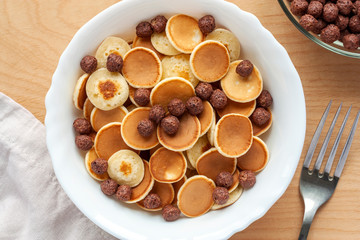 Trendy food during coronavirus quarantine - pancake cereal. Mini pancakes in white bowl with cocoa crispy balls on wooden table. Breakfast for kids, tiny pancakes.