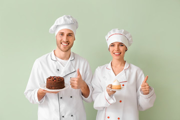 Young confectioners with tasty desserts showing thumb-up gesture on color background