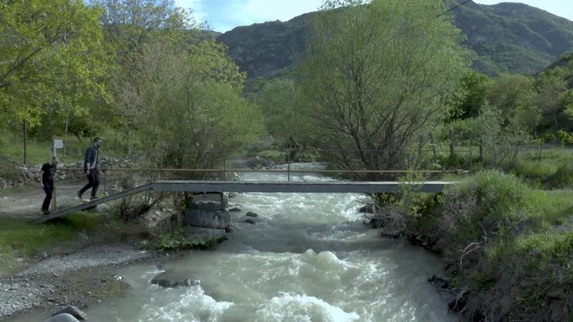 Two Friends Crossing On The Small Metal Bridge Over The River With Rapid Water In Kaspi, Georgia On A Beautiful Day. - crane shot