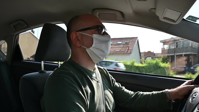 Turin, Piedmont, Italy. May 2020. Coronavirus pandemic: portrait of a Caucasian man driving a car wearing a white mask. He is traveling straight on a suburban road.