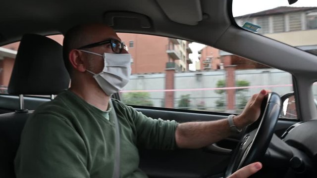Turin, Piedmont, Italy. May 2020. Coronavirus pandemic: portrait of a Caucasian man driving a car wearing a white mask. He makes a turn and then parks next to other cars.