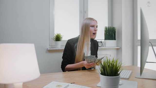 Elegant blond female sitting at desk at workplace with computer holding in hands bundle of cash looking astonished at monitor, and throwing money in air and happily making yes gesture.