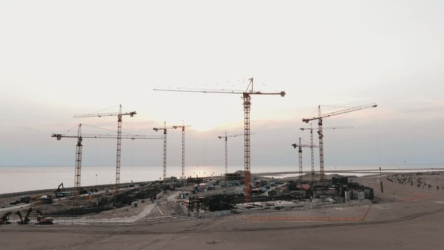 beautiful aerial drone flying at a construction site, a truck is driving along the road, a crane raises a wall against the backdrop of a sunset on a bay shore slow motion