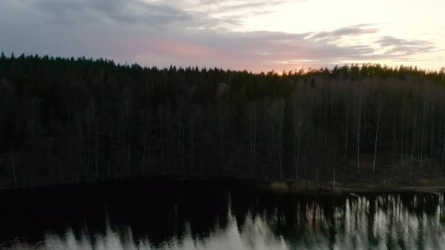 Flying over a lake at sunset a drone rises above the surrounding forest to reveal the sun just as itis dipping below the horizon.