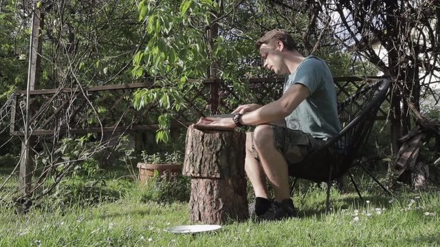 Young man sits on black chair in sunny backyard garden next to a wooden  construction, with hanging green plant of it, slicing meat for BBQ. wide shot 29.97 fps. trucking shot