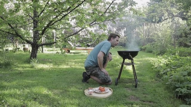 Young man in a beautiful quiet green and sunny backyard garden preparing BBQ. He is blowing air into grill with mouth and lungs to start fire. One person. wide shot barbecue 29.97 fps