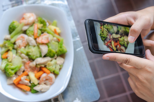 Closeup of smartphone screen with young woman fingers take a photo of food, blurred mixed vegetables with shrimps and chicken. Phone photography of lunch or dinner for social networks.