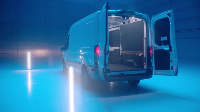 Track around rear of white cargo vans in an empty warehouse with its rear doors open lit in blue