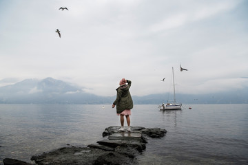 Fototapeta na wymiar A girl in a green jacket and a pink hat stands with her back on the lake and enjoys the view. Spectacular view of Lake Como. Cloudy and rainy weather, seagulls fly in the sky
