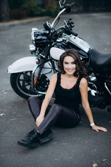 Fototapeta na wymiar Young beautiful biker woman in a black tank top and leather pants sits on the pavement near a motorcycle. The concept of speed and freedom. Soft selective focus.