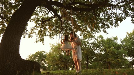 Mom shakes a healthy little daughter on swing under tree in sun. mother plays with child they are swinging on rope on an oak branch in forest. girl laughs, rejoices. Free family having fun in park