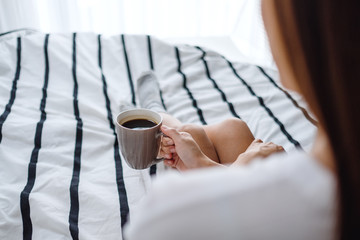 A beautiful woman drinking hot coffee in a white cozy bed at home in the morning