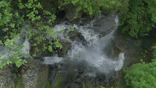 The drone floating over a waterfall we see the water falling down into a clear stream shaded by green bushes and grass.