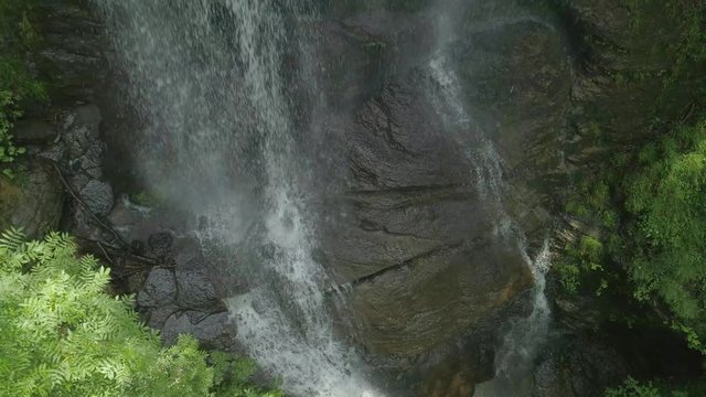 A closeup view of waters hitting rocks with the drone pulling back into the sky through vibrant green leaves.