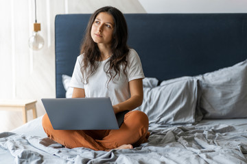 Beautiful young brunette girl with a laptop sitting on the bed. Stylish modern interior. A cozy workplace. Shopping on the Internet