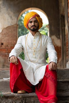 Portrait of an young Indian man in ethnic wear with turban sitting in front of an old vintage house.