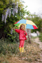 A girl of three years old in a pink jacket, rubber boots and with a multi-colored umbrella walks alone in a green spring park in the open air.