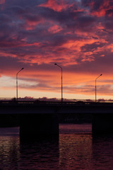 Wonderful sunset sky with colorful clouds and sunbeam over black bridge over river with square frame with reflection of sunlight in water, minimalist landscape.