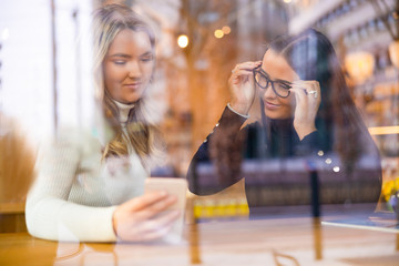 Two Beautiful Women Looking At Phone Together In Cafe Seen Through Window