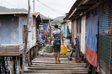 unrecognizable young man carrying jerrycans filled with water walking over wooden path over the...