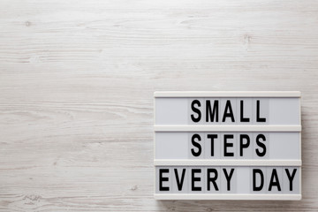 'Small steps every day' words on a lightbox on a white wooden surface, top view. Overhead, from above, flat lay.