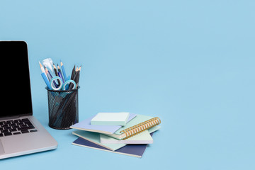Laptop, stack of notepads, stand with stationery on a blue pastel background. Student workspace concept.
