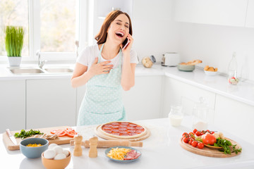 Obraz na płótnie Canvas Photo of beautiful cheerful housewife enjoy hobby cooking family recipe dinner pizza speaking telephone laughing out loud wear t-shirt apron standing modern kitchen indoors