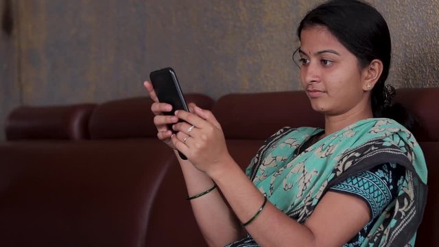 Girl in casual dress busy on mobile while sitting on sofa at home - concept of Indian woman using social media, Internet, digital divide and technology at home.