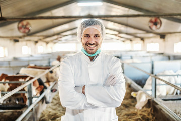 Cheerful caucasian veterinarian in protective white uniform with hair net on head standing in barn with arms crossed.