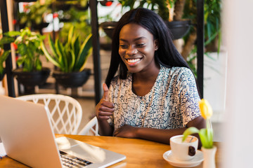 Portrait of an african american young woman with thumbs up sitting at cafe with laptop writing her blog
