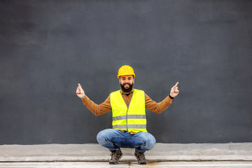 Young smiling attractive constructor crouching in front of gray wall and showing up with his...