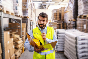 Young smiling employee in vest holding folder with documents and checking out inventory.