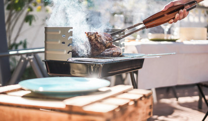 Chef grill t-bone steak at barbecue sunday lunch outdoor - Man cooking meat for a family bbq meal outside in backyard garden - Summe lifestyle and bbq food concept - Focus on tongs, hand