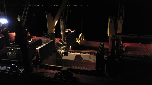 Close-up cargo bulker with coarse grain in holds. Night at merchant vessel. Ship crane lower telescopic handler with ladle in vessel storage with agricultural crops. Merchant boat work at nighttime