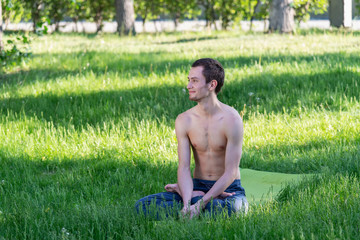 A young, beautiful white man in sports clothes is doing a stretch in the Park on the green grass. Prepare a warm-up for yoga or fitness classes. violation of isolation mode