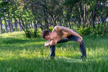 A young, beautiful white man in sports clothes is doing a stretch in the Park on the green grass. Prepare a warm-up for yoga or fitness classes. violation of isolation mode