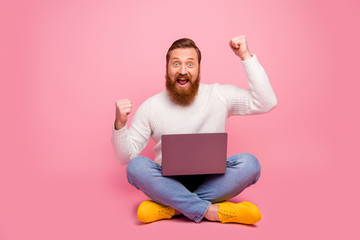 Full length photo excited delighted guy sit floor legs crossed folded work computer celebrate...