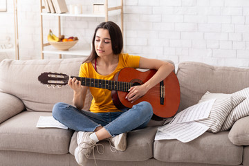 Stay home hobbies. Pretty millennial woman playing acoustic guitar on sofa in living room