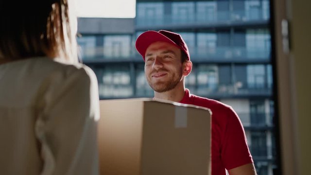 Cheerful handsome postman in red uniform delivering big cardboard box to beautiful customer young woman in modern business district. Home order delivery, transportation.