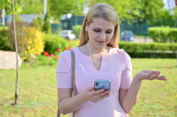 Portrait of a cute caucasian girl outdoors talking on a smartphone. Pretty blonde model posing while standing in sunny weather in a city park.