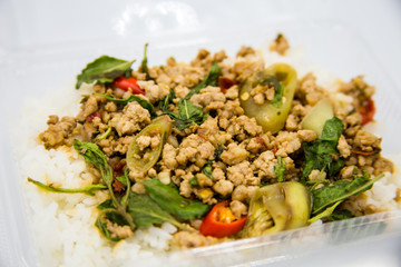 Rice topped with stir-fried pork and basil in a box placed on a white ground.