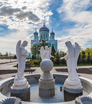 Sculpture of three angels on the background of the Orthodox Church and blue sky with clouds in summer