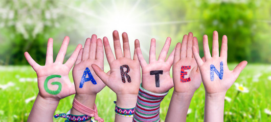 Children Hands Building Colorful German Word Garten Means Garden. Sunny Green Grass Meadow As Background