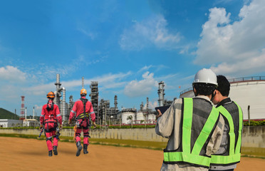 Team Worker in Oil Refinery discussion for inspection by hand holding tablet on power plant with refinery plant structure and two worker wearing safety harness walking in background.