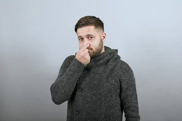 Young Bearded Dark Haired Man In Black Stylish Shirt On White Background, Disappointed Male Unpleasant, Covered Nose With His Hand From Bad Smell. The Concept Of People Who Stink