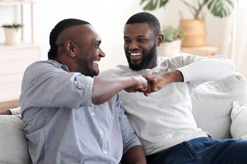 Father-Son Friendship. Black Millennial Guy Bumping Fists With Senior Dad At Home