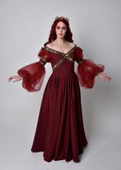 Portrait of a beautiful woman with red hair wearing  a  flowing Burgundy fantasy gown and golden crown.  full length standing pose, isolated against a studio background
